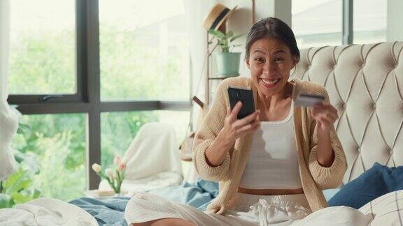 快乐的亚洲女孩大学穿着白色的奶油睡衣躺在蓬松舒适的床上使用信用卡网上购物成功支付手机兴奋看着相机在卧室的家
