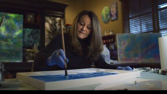 在室内艺术工作室里一名60多岁的女白种艺术家用画笔蘸上白色油画颜料并将其涂在画布上