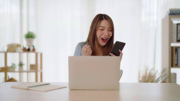 有趣的欣快的年轻亚洲妇女庆祝赢得或获得电子商务购物优惠的电脑笔记本电脑兴奋快乐的女孩赢家看着笔记本庆祝成功