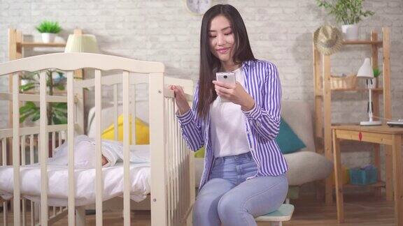一名年轻的亚洲女性拿着智能手机在客厅的婴儿床里抱着一个婴儿