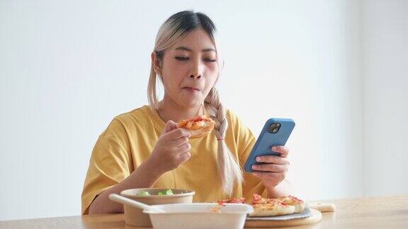 年轻的亚洲女子愉快地吃着披萨在家里用手机亚洲休闲用餐