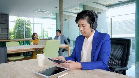 画像的亚洲女商人工作与智能手机和戴无线耳机而坐在现代办公室