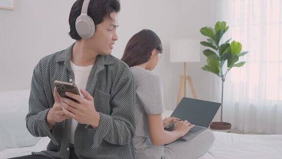 年轻的亚洲夫妇坐在床上微笑着男人戴着耳机女人用着笔记本电脑年轻夫妇一起躺在床上听音乐玩笔记本电脑