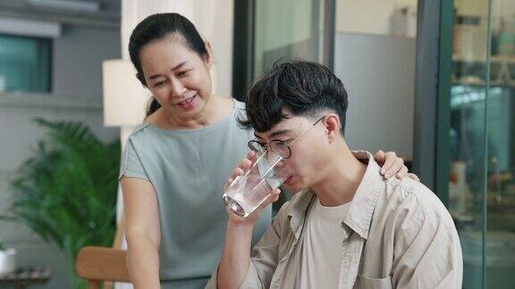 年轻的亚洲男子喝水他的母亲看着他的作品