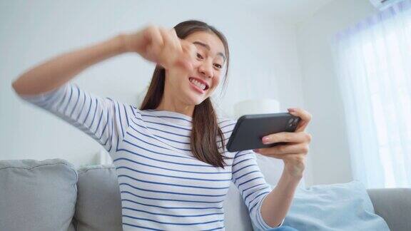 亚洲年轻漂亮的女子在家里玩智能手机游戏迷人的休闲女孩感到快乐和放松坐在沙发上玩着玩着手机屏幕播放视频享受胜利在家里