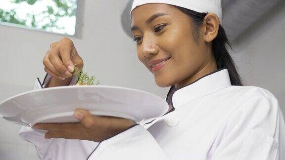 在专业厨房里美丽的亚洲女厨师俯身装饰盘子里的食物看起来很开心