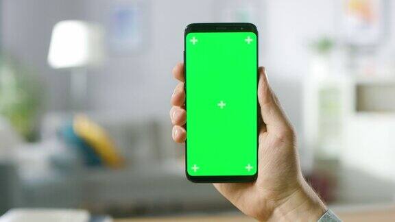 一个男人的手握绿色模拟屏幕智能手机的特写现代移动电话在舒适的客厅或家庭办公室的背景