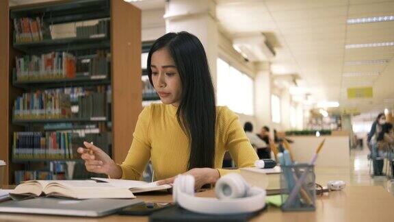 亚洲学生女孩坐在桌边一边看书一边做作业用笔记本电脑在国外通过网友联系打视频电话在图书馆里开心地笑着