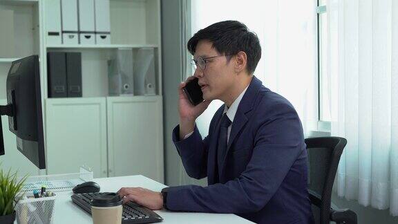 严肃的亚洲商人穿着西装在办公桌上使用电脑被手机打扰他在办公室里心烦意乱忙得不可开交