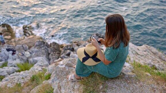 SLOMO女游客坐在岩石边缘在海边用智能手机拍照