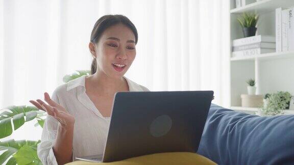年轻的亚洲女性坐在家里客厅的沙发上用笔记本电脑与顾客进行视频通话