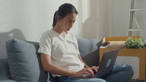 年轻的亚洲女性搬回家后坐在沙发上使用笔记本电脑在家工作