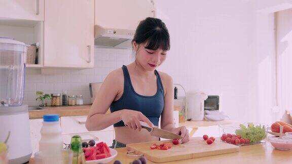 一个穿着运动服的亚洲女人开心地切蔬菜把西红柿放在玻璃碗里做沙拉她在厨房里拥抱快乐和健康的生活方式通过营养丰富的烹饪促进健康