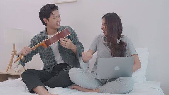 年轻迷人的亚洲夫妇坐在一起看笔记本电脑在卧室里男人和女人在家里的床上用笔记本电脑在线弹吉他爱情和夫妻关系