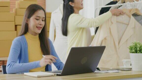 年轻的亚洲时尚企业家在餐桌上用笔记本电脑查看订单而她的母亲则在准备在家办公时出售的衣服