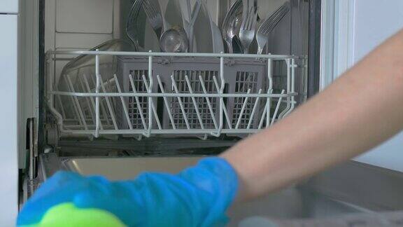 内置洗碗机银色餐具放置在一个托盘和碗在较低的抽屉车门是用浅绿色的餐巾擦的一只手戴着蓝色的橡胶手套特写镜头