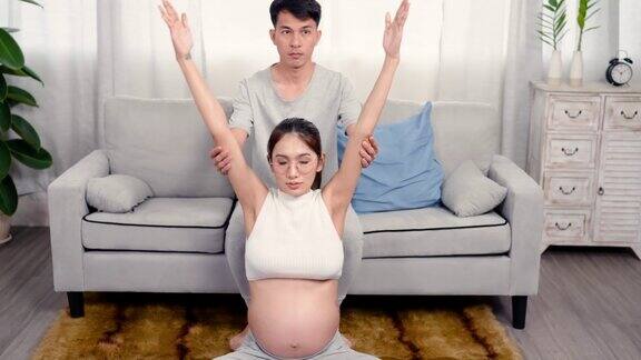 4k怀孕的亚洲妇女坐在一个放松的瑜伽姿势坐在客厅中间摇摆前后手臂交替举起与丈夫挽臂在背后保护安全