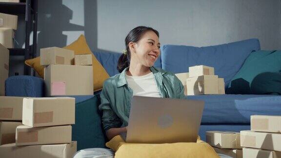 年轻的亚洲女售货员在网上商店使用笔记本电脑准备包裹发货给客户在家里