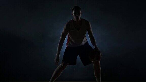 在一个黑暗的房间里一个篮球运动员在球场上运球在烟雾中有一个慢动作的背光