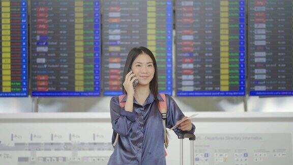 年轻的亚洲女子手持护照和登机牌站在航空公司的时刻表板前在机场讲电话专业或讨论信息