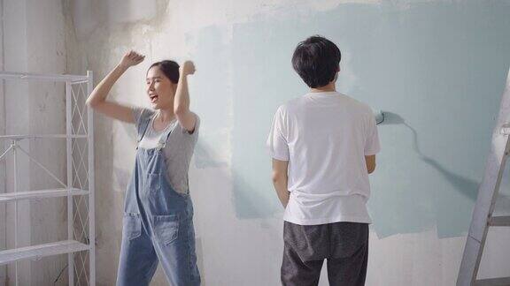 年轻的亚洲夫妇在他们的新公寓装修期间一边用滚轴修理和刷蓝色油漆的墙壁一边跳舞