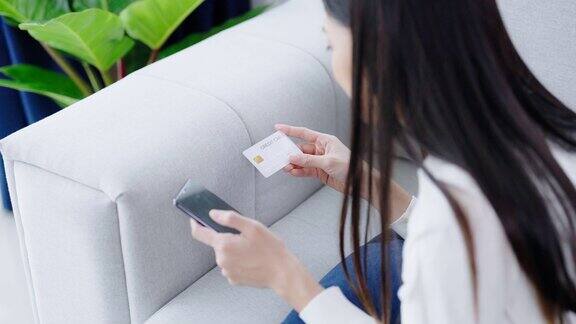 年轻的亚洲女子躺在客厅的沙发上用智能手机上的银行卡通过互联网进行网上银行支付用信用卡在手机上网购