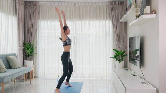 一名亚洲年轻女子在家中客厅的智能电视上一边观看视频一边进行高强度的有氧健身、体重锻炼、跳杰克运动