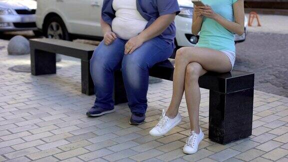 美女用智能手机转开胖人坐在长椅上形成对比