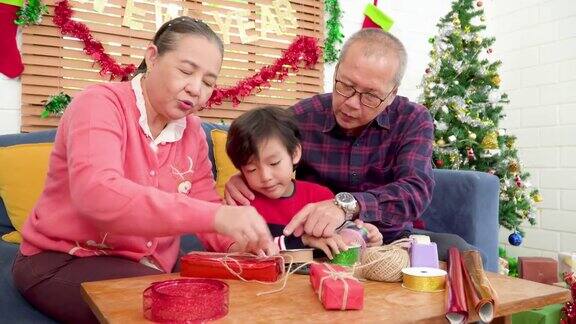 亚洲爷爷帮助奶奶在沙发上为圣诞节庆祝包装礼物而一个男孩在他的手臂感兴趣想帮助装饰圣诞节的家