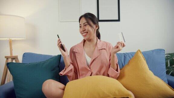 快乐年轻的亚洲女士使用手机订购网上购物产品用信用卡支付账单坐在沙发上客厅在房子