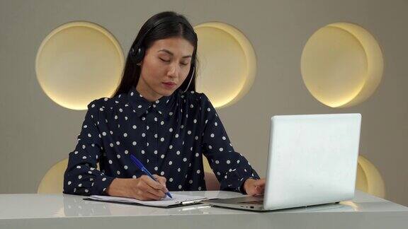一位年轻的亚洲女性戴着耳机看着笔记本电脑屏幕听和学习在线课程