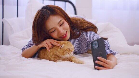 亚洲美女在家里用手机和家猫拍照年轻漂亮的女主人躺在床上心情愉快放松与宠物在卧室度过闲暇时光
