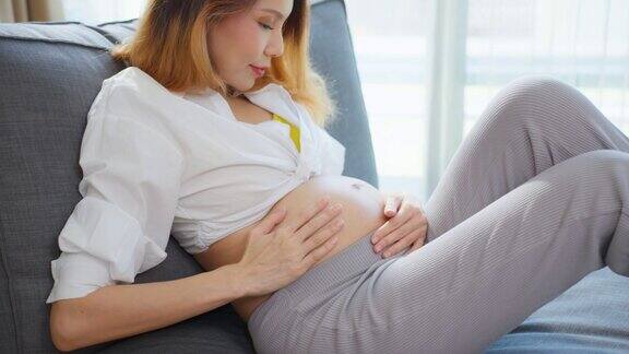 亚洲年轻美丽的孕妇抱着并抚摸她的肚子迷人的准孕妇妈妈坐在室内客厅的沙发上看着肚子幸福和爱