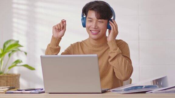 企业家英俊的商业亚洲人穿休闲衬衫和耳机听有乐趣的音乐快乐的自由职业者在休息时戴着无线耳机在家里的桌子上跳舞