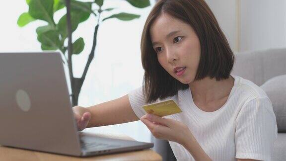 年轻的亚洲女性手拿信用卡在家里客厅的笔记本电脑网上购物4K分辨率通过笔记本电脑银行
