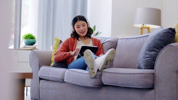 平板电脑耳机和女人在沙发上放松音乐流媒体应用程序社交媒体和在家看视频z世代亚洲人或年轻的韩国女孩坐在沙发上数码科技和听音频