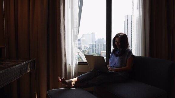 剪影亚洲妇女使用笔记本电脑在豪华家庭客厅的大窗户享受工作网上购物检查社交网络阅读新闻与笔记本电脑在现代椅子