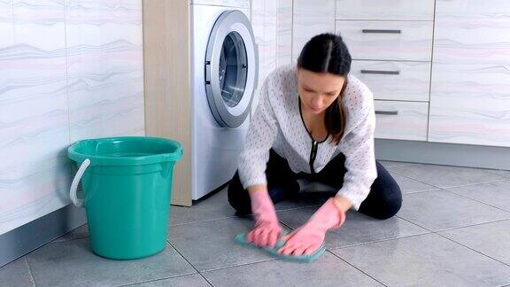 戴着粉色橡胶手套的疲惫的女人用一块布洗厨房的地板看着镜头