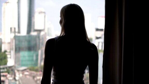 女人揭开窗帘向窗外望去的剪影城市摩天大楼景观