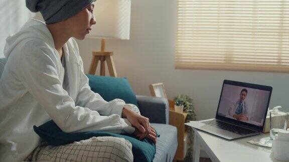 年轻的亚洲妇女生病的癌症使用笔记本电脑交谈医生坐在沙发上的客厅在家里