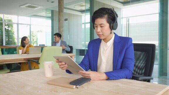 画像的亚洲女商人工作与数字平板电脑和戴着无线耳机而坐在现代办公室商务人士同事坐在背景