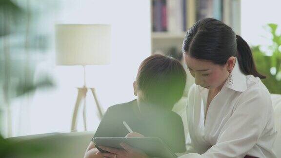 多莉展示了一个富有的亚洲妇女和她的儿子在客厅的数字平板电脑上一起画画开心的微笑
