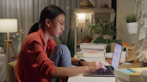 年轻的亚洲女性穿着休闲拿着笔记本电脑坐在桌子前晚上在家里的客厅里思考创作工作想法居家办公
