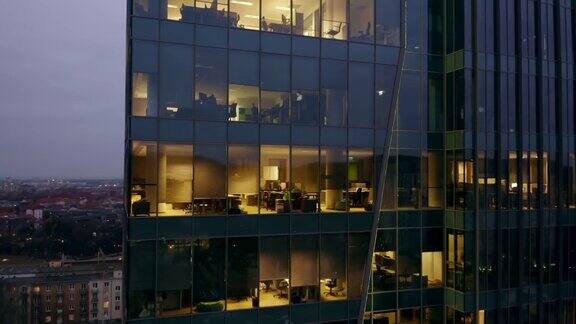 一架无人机击中了一座夜间的摩天大楼那里有办公室和工作到很晚的人他们解决业务问题坐在电脑前无人机绕着玻璃摩天大楼从右到左飞行