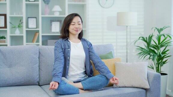 年轻的成年亚洲女子闭着眼睛坐在客厅的沙发上冥想