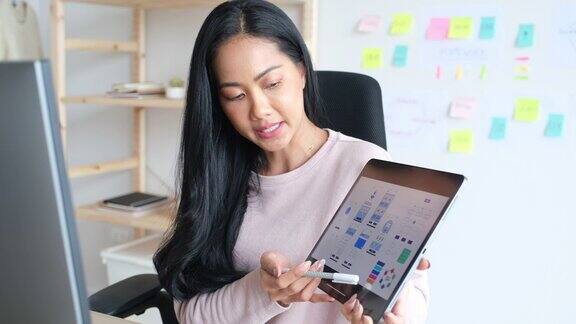 年轻的ui设计开发人员女程序员手持数字平板电脑屏幕上显示着移动应用程序原型同时与团队在家庭办公室进行虚拟视频会议在家工作
