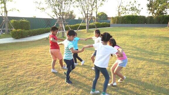 在阳光明媚的夏日里一群快乐的亚洲微笑的幼儿园小朋友手拉手嬉戏跳舞在公园里的绿草地上打转、站成一圈