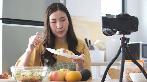 年轻的亚洲女性美食博主穿着围裙在她的美食频道上录制鸡蛋火腿配米饭的视频女厨师在自家厨房用数码相机播放视频向大家讲解煎蛋卷和火腿配米饭