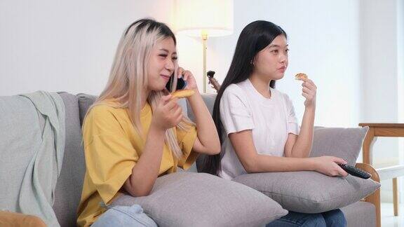 两个年轻的亚洲女性朋友兴高采烈地吃着披萨一边打手机一边看电视坐在家里客厅的沙发上亚洲休闲用餐
