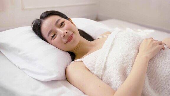 美丽性感的亚洲女性的肖像与迷人的微笑享受新鲜柔软的床上用品亚麻床垫在白色的卧室现代公寓可爱的亚洲女人睡觉休息晚安睡概念